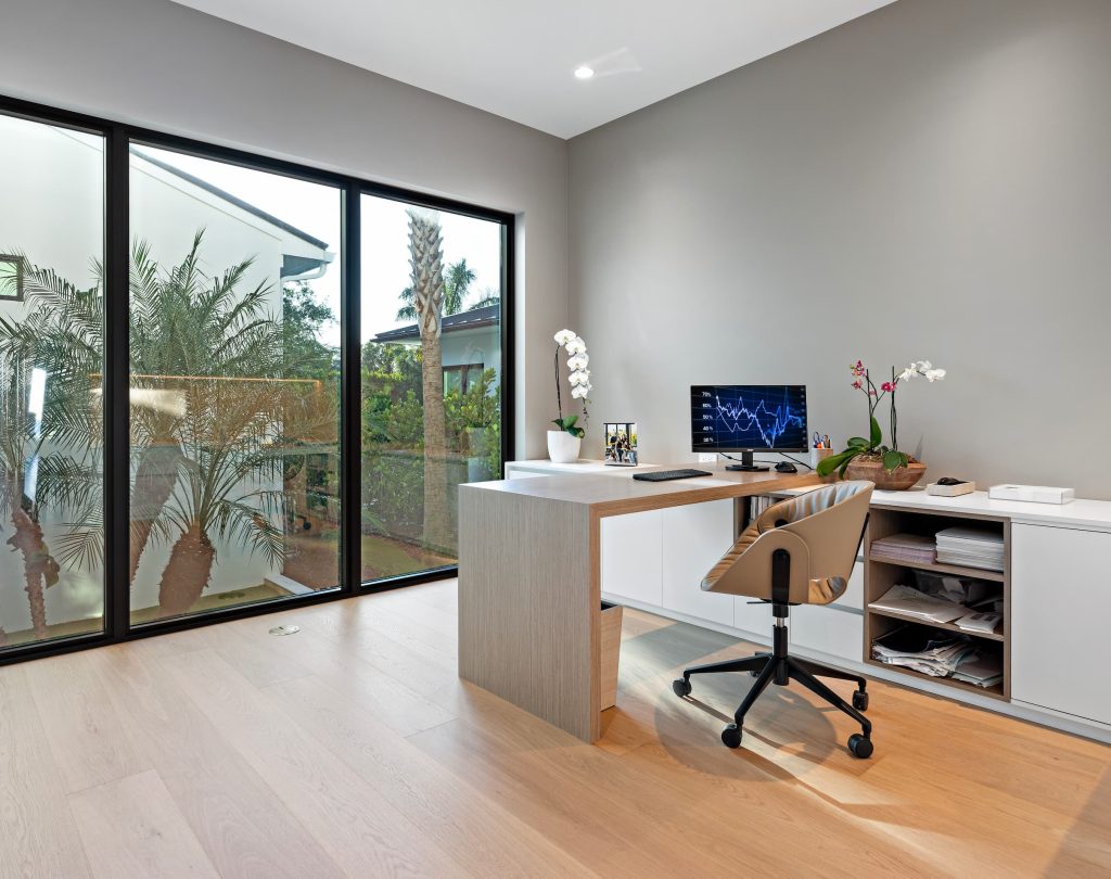 Minimalist home office featuring large black-framed windows, a modern desk setup, wood flooring, and natural light with views of tropical landscaping