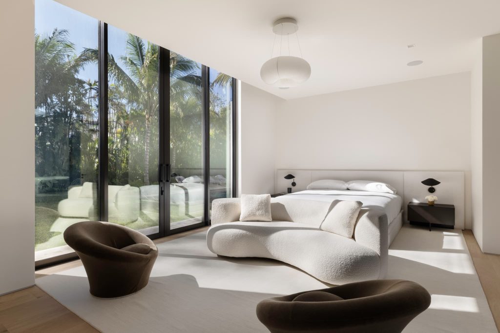 Modern bedroom featuring floor-to-ceiling glass doors, minimalist furniture, soft neutral tones, and abundant natural light with views of a tropical garden