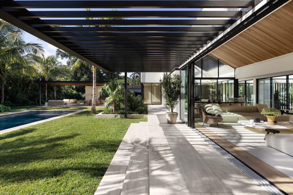 Modern indoor-outdoor living area featuring a shaded pergola, expansive sliding glass walls, contemporary seating, and seamless access to a landscaped garden and pool