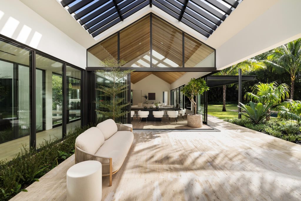 Modern indoor-outdoor living space featuring expansive glass walls, a skylight roof with wood ceiling, contemporary seating, and seamless connection to a lush tropical garden