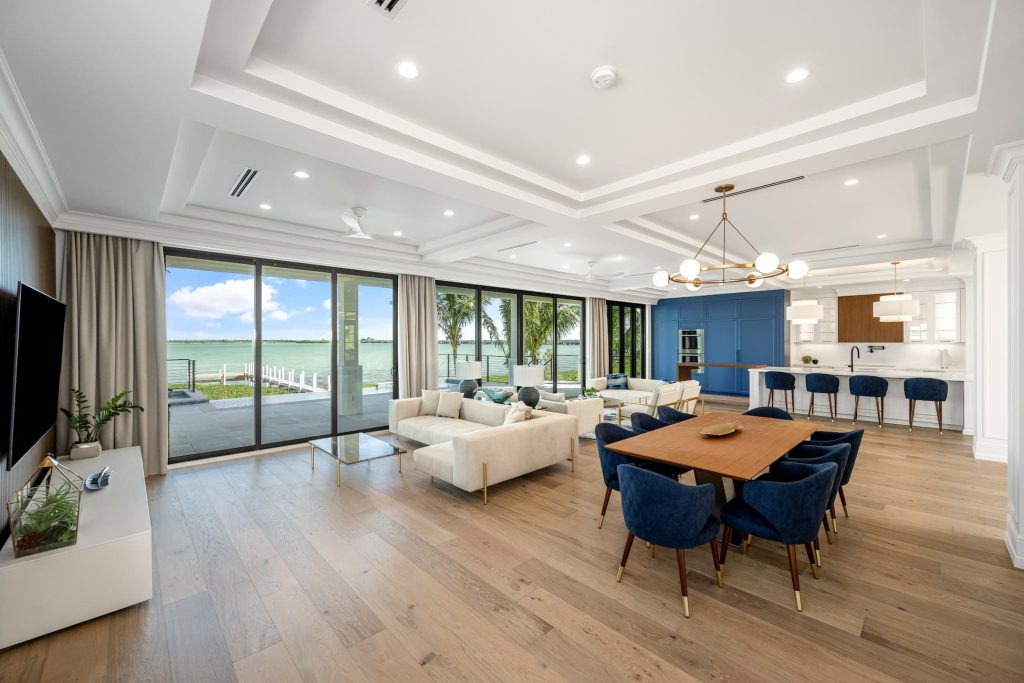 Open-concept waterfront living and dining area featuring large sliding glass doors, modern furnishings, wood flooring, and seamless indoor-outdoor access to a terrace overlooking the water